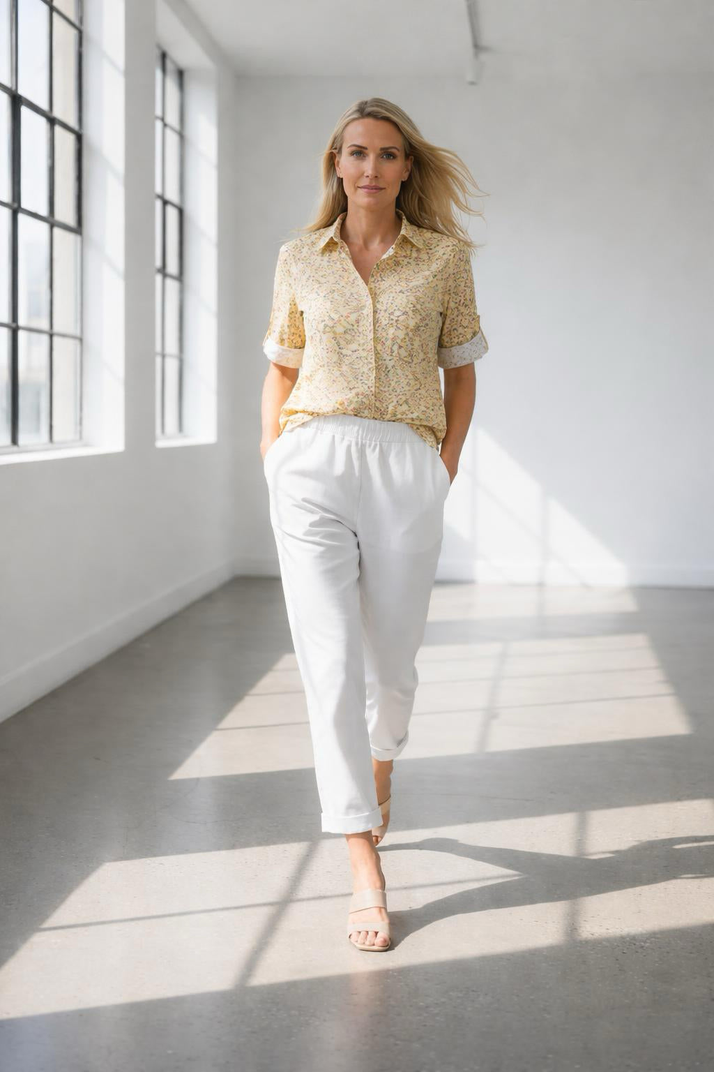Woman in a yellow patterned shirt and white pants standing in a bright room with large windows.