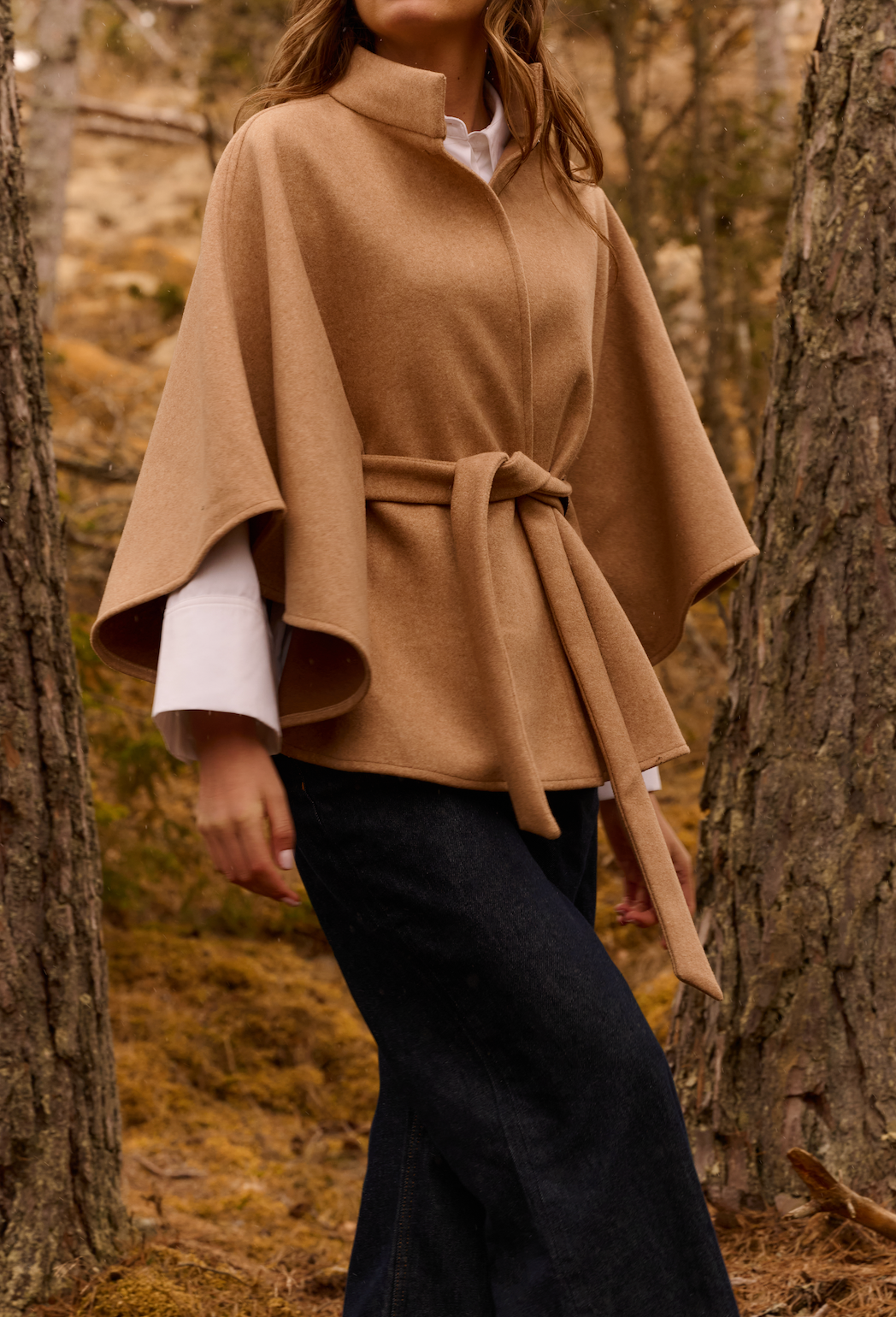 Model wearing the YAYA Soft Woven Poncho with Collar & Belt in camel, styled with a white shirt and dark denim in an autumn woodland setting.